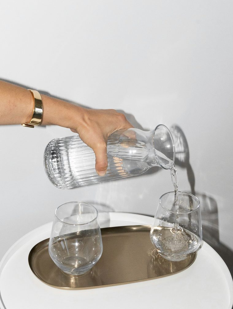 Una mano sirve agua desde una jarra de cristal acanalado a dos vasos sobre una bandeja