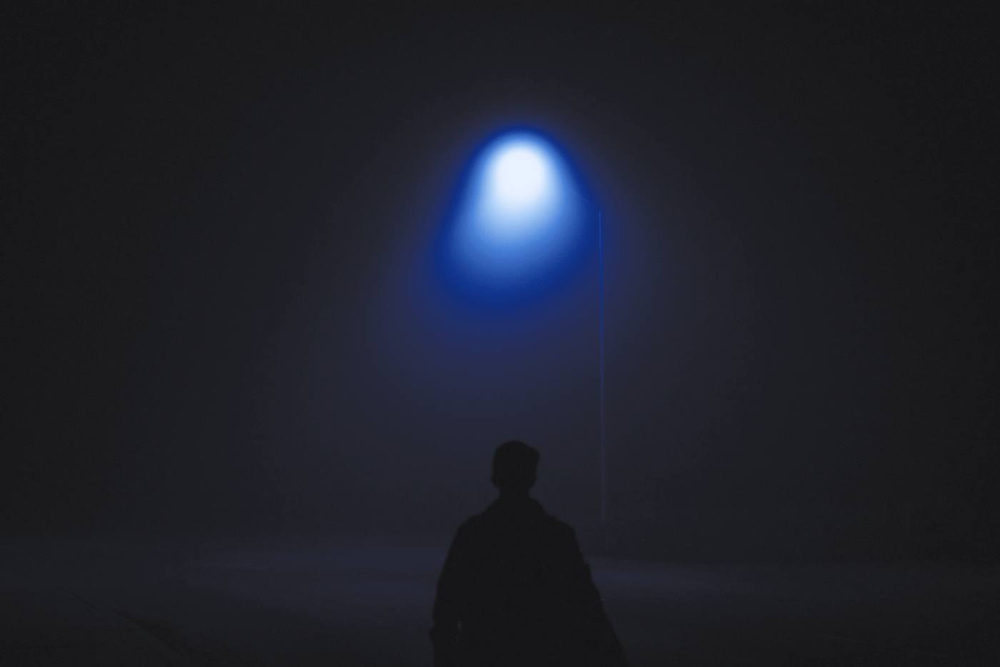 La silueta de una persona de espaldas bajo una farola entre la niebla, en tonos de azul