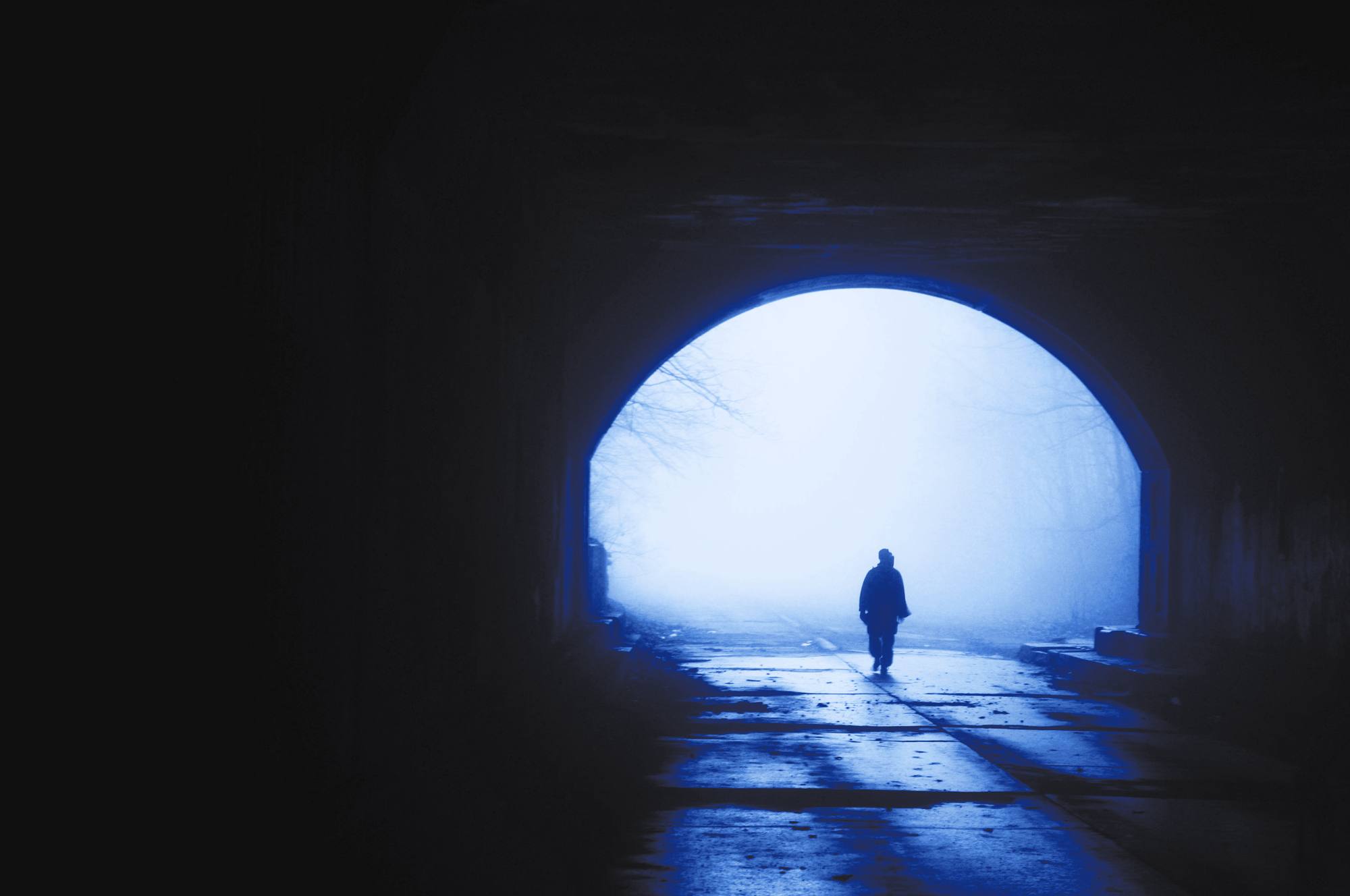 Una figura camina hacia la salida de un túnel inundada de niebla, en tonos de azul