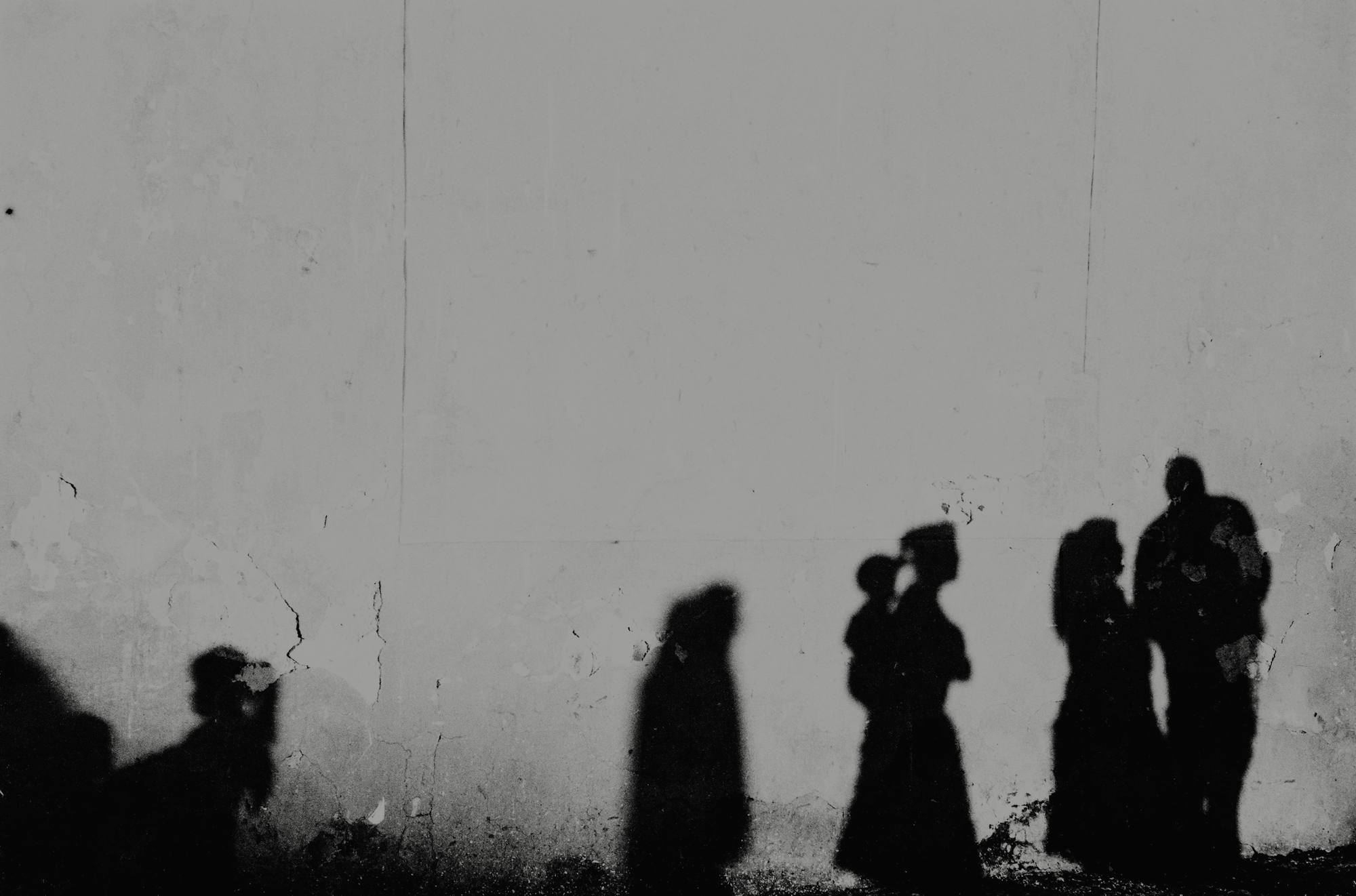 Sombras de un grupo de personas proyectadas sobre un muro blanco descascarado, en blanco y negro