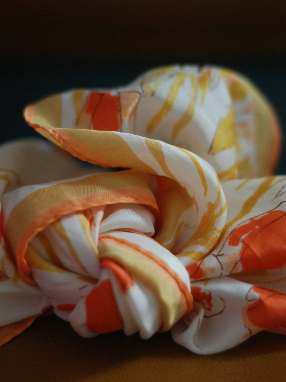 Pañuelo de seda estampado en naranja y marfil, anudado sobre una mesa con fondo oscuro