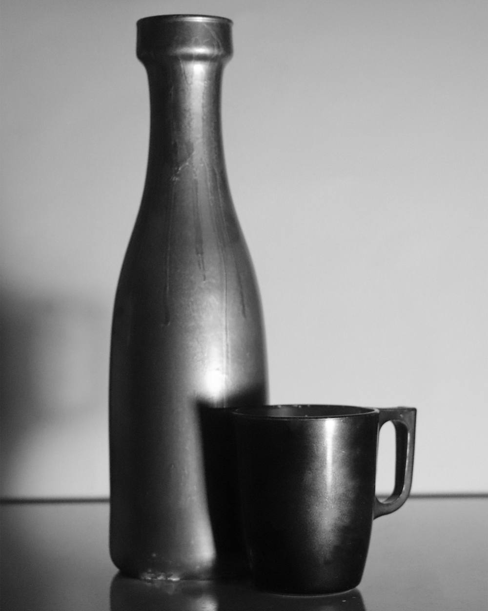 Botella y taza metálicas sobre una mesa que las refleja, en blanco y negro