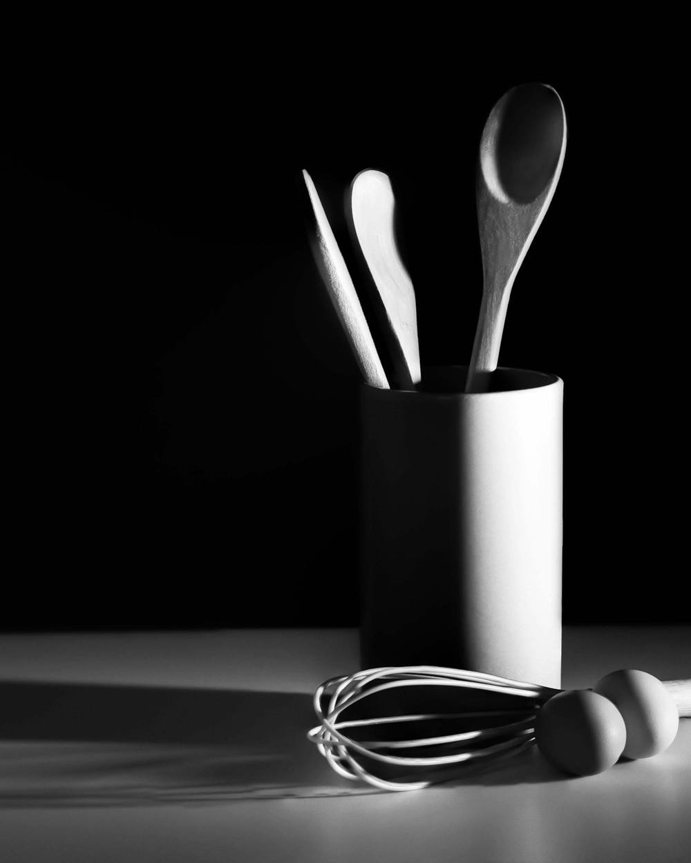 Cuchara de madera, espátulas y batidor sobre una mesa a contraluz, en blanco y negro