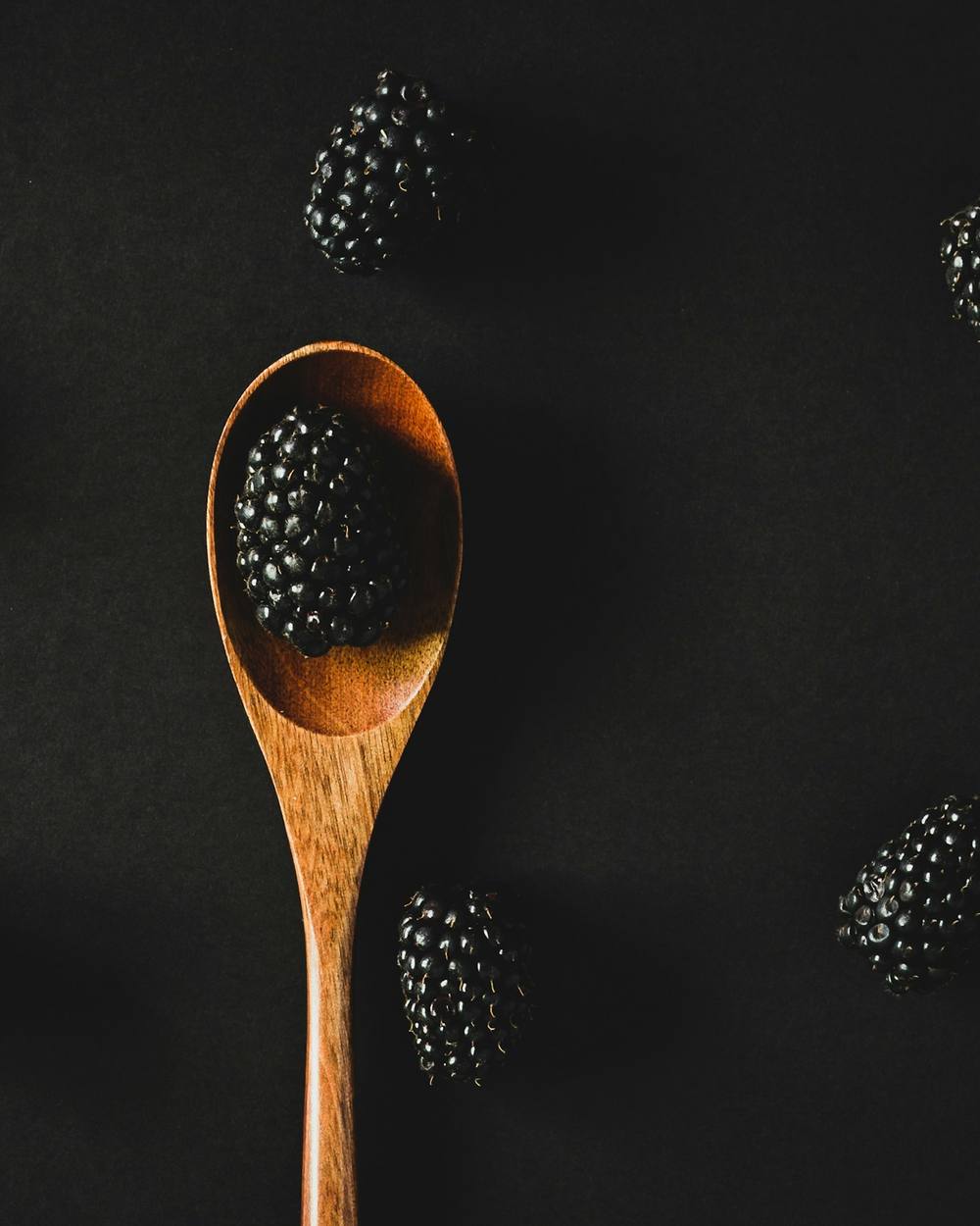 Una cuchara de madera con una mora, rodeada de moras sueltas sobre fondo negro
