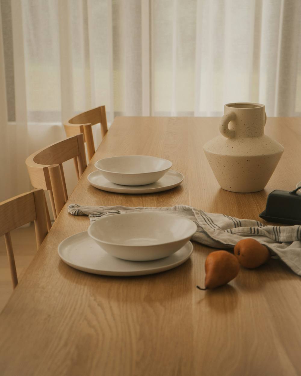 Mesa de madera con vajilla clara, un jarrón y peras junto a una cortina