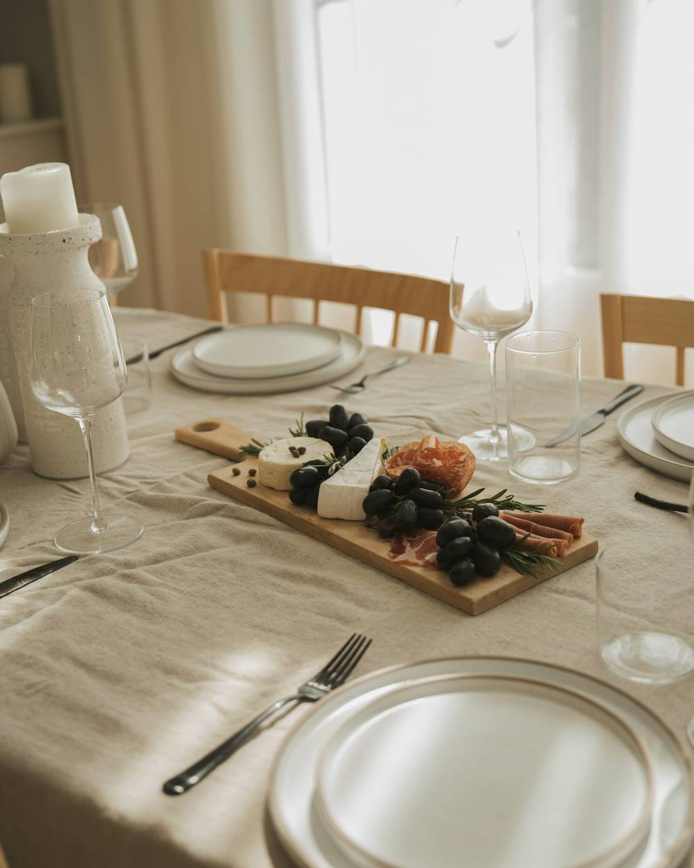 Mesa de lino con platos blancos, una tabla de quesos y vasos junto a la ventana