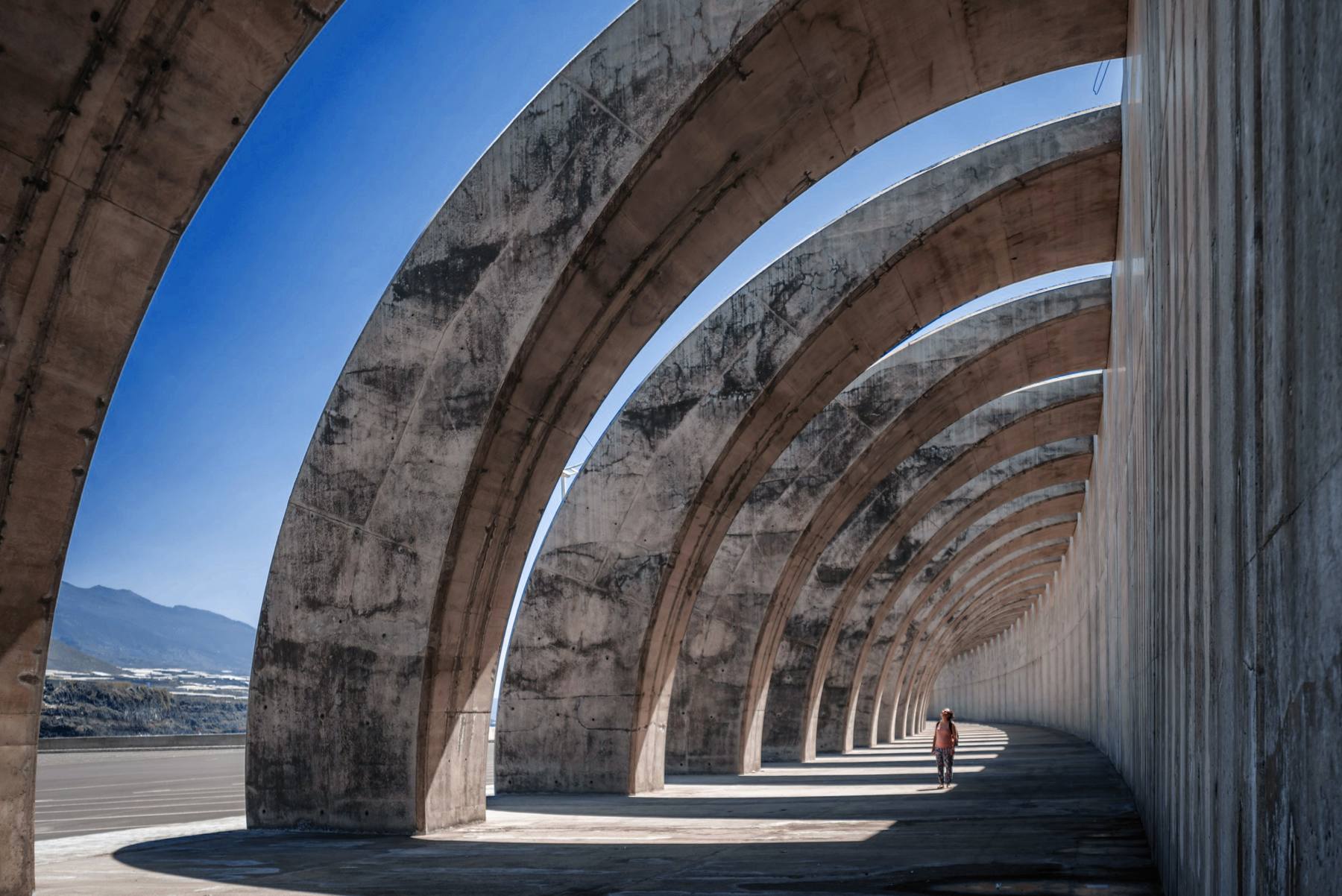 Arcos de hormigón que se suceden a lo largo de un paseo, con el cielo azul asomando entre ellos