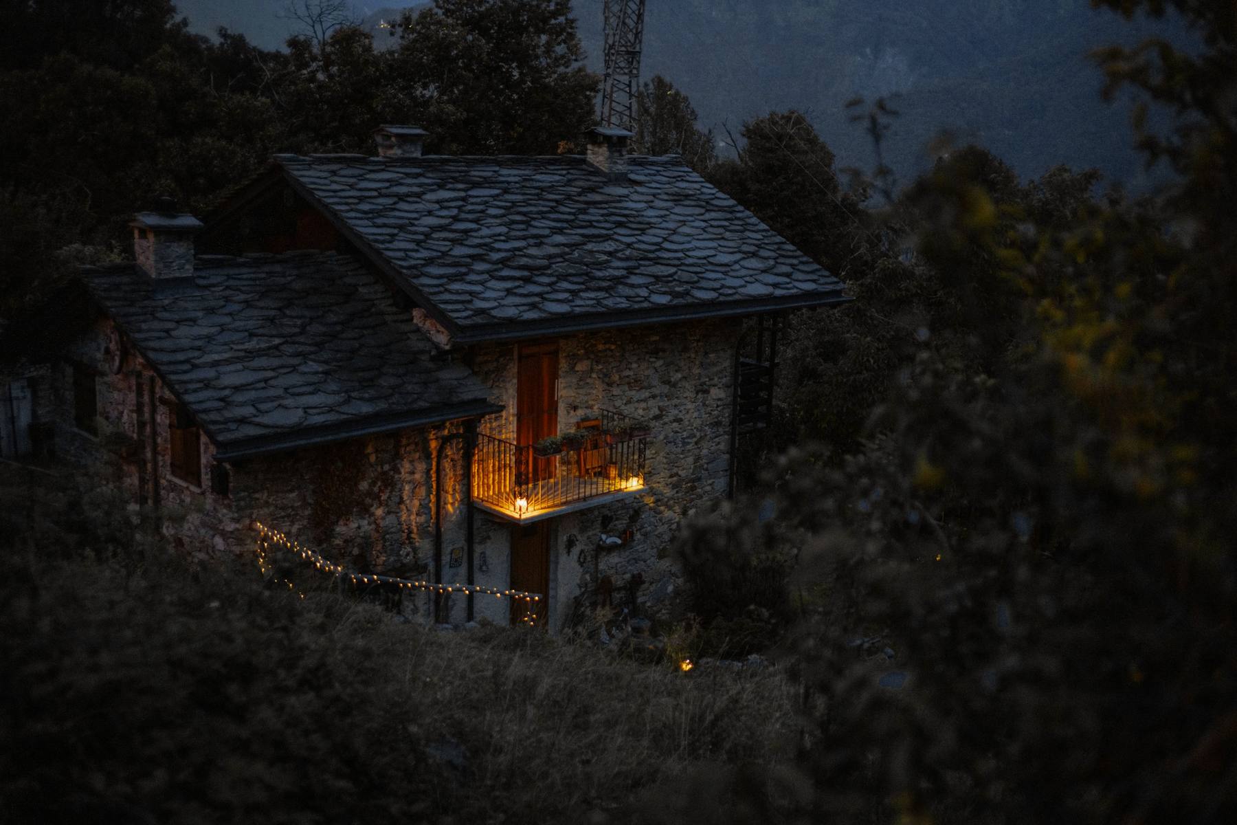 Una casa de piedra con el techo de pizarra y una ventana encendida, entre árboles oscuros en la ladera al caer la tarde