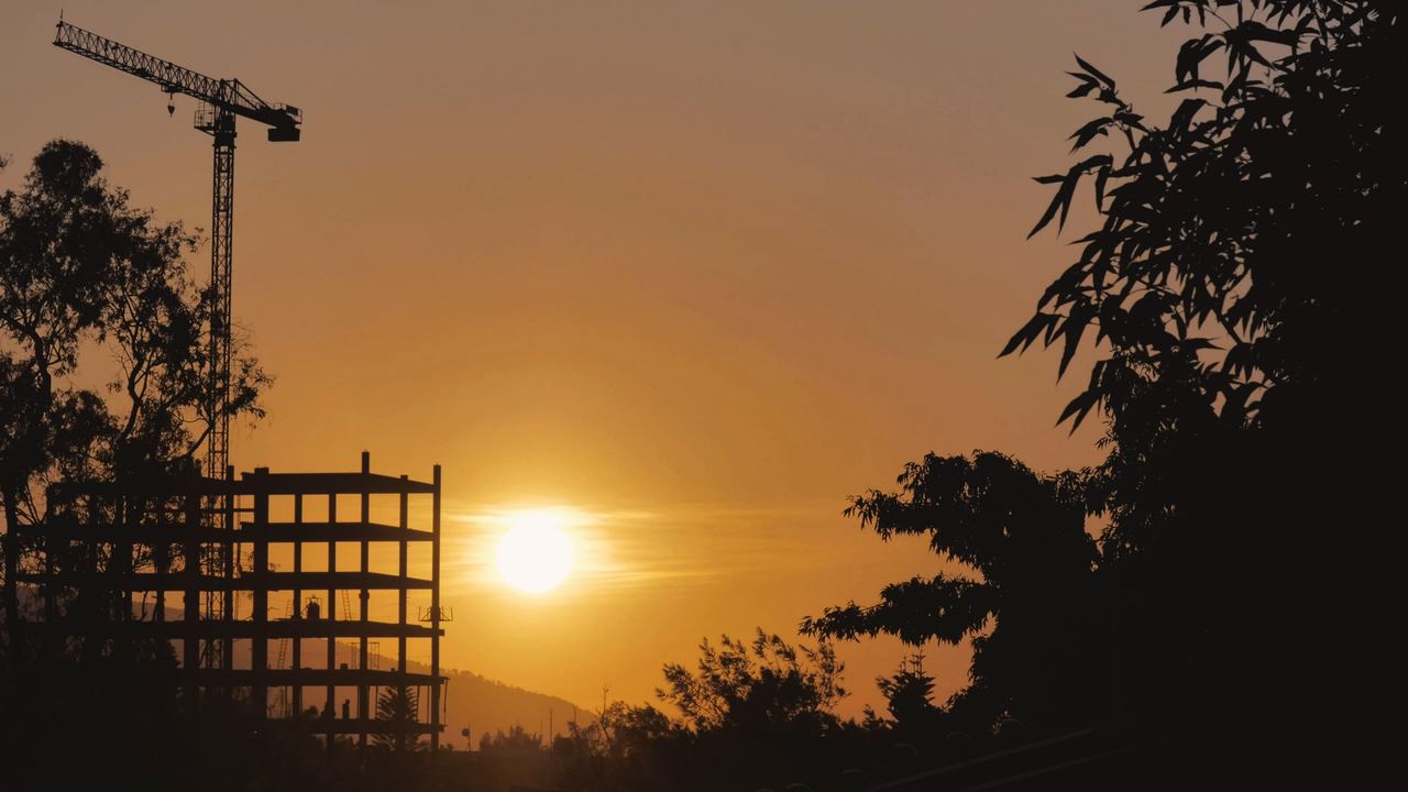 Estructura de un edificio en construcción con una grúa, recortada contra un sol que se pone entre los árboles