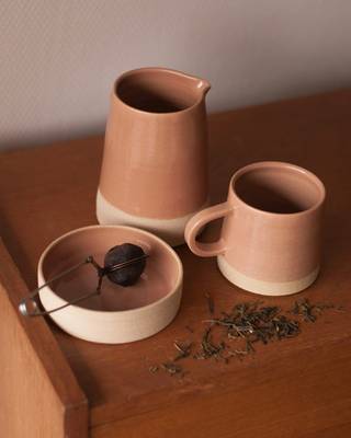 Jarra y taza de barro rosado junto a un cuenco, sobre una mesa de madera