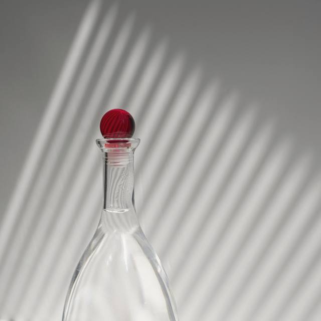 Una garrafa de vidrio con tapón rojo y sombras de persiana