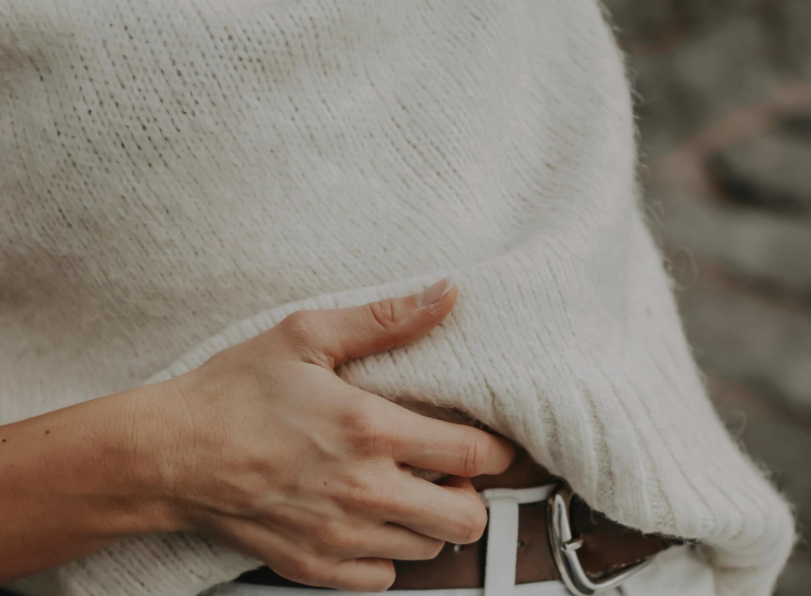 El torso de una persona con un suéter claro de punto, la mano en el cinturón de unos jeans blancos