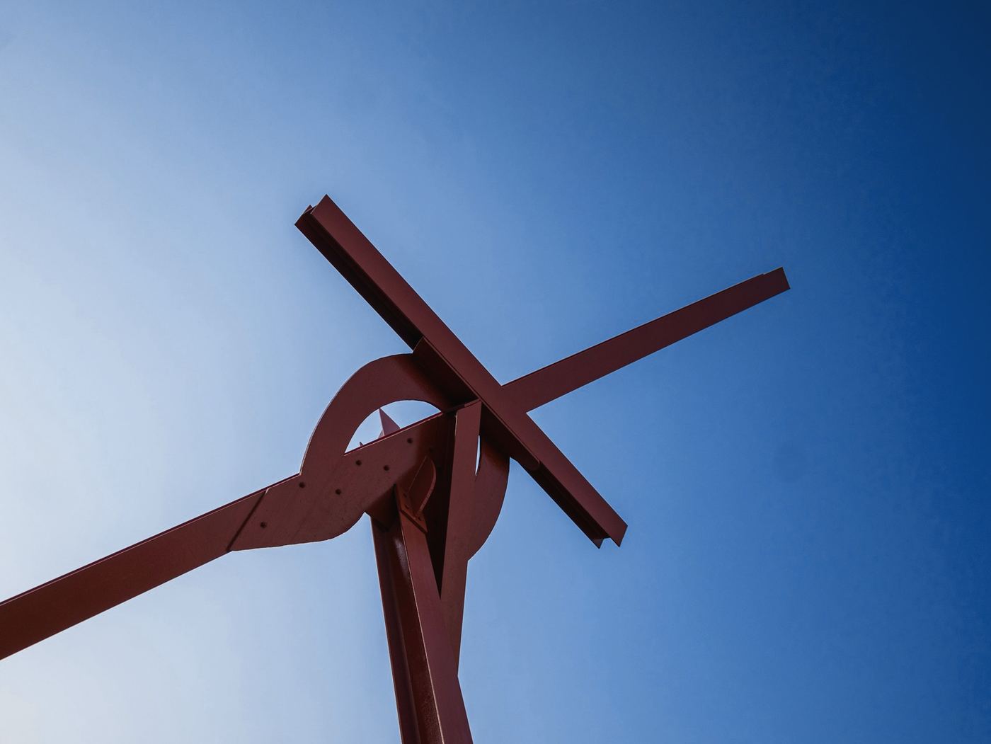 Escultura de vigas de acero rojizo en cruz sobre un cielo azul