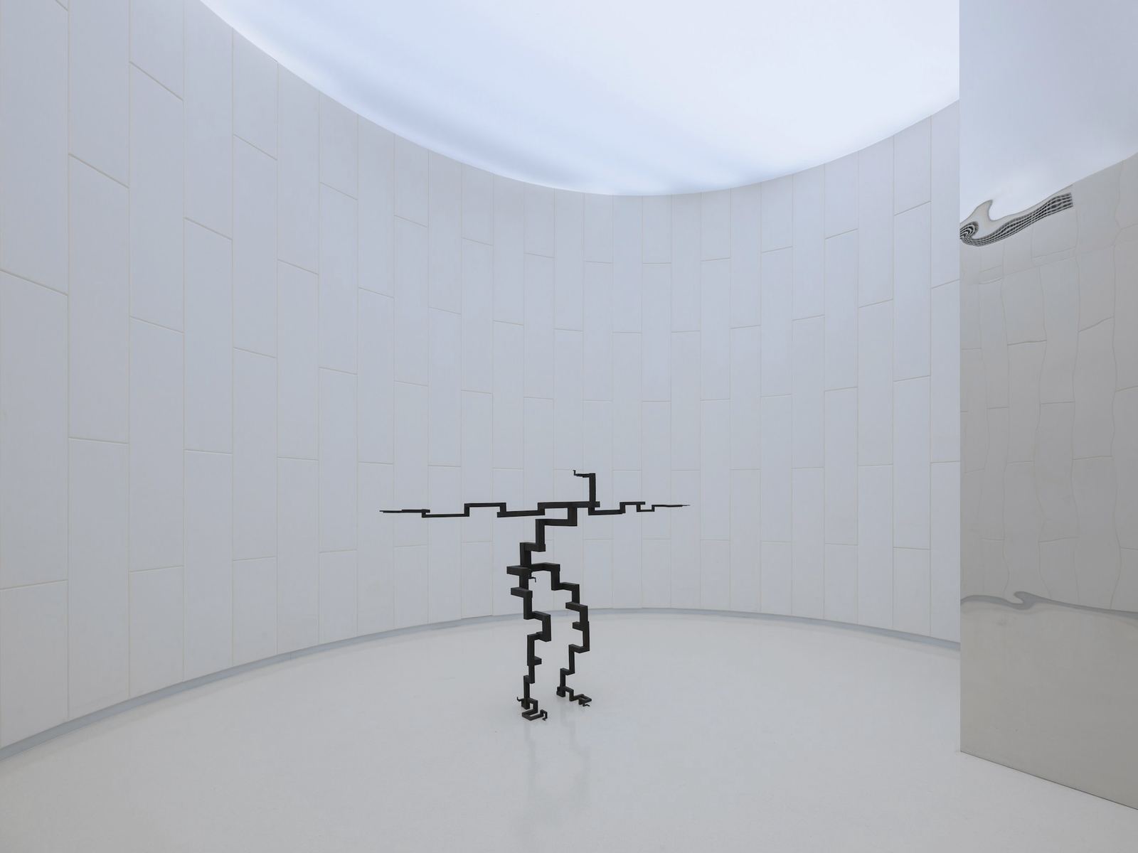 Sala circular blanca con una escultura de acero oscuro en el centro y un espejo curvo a un costado