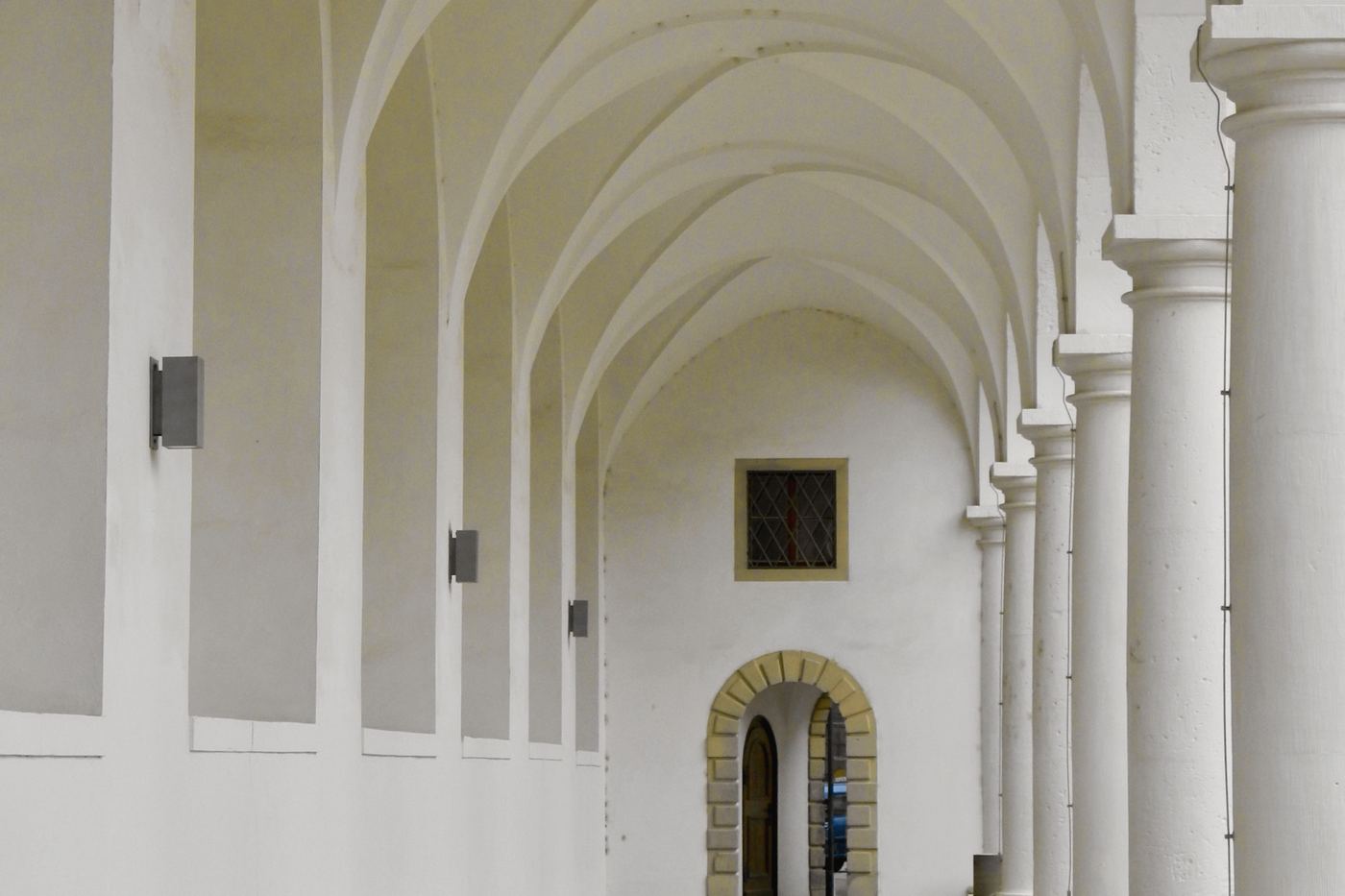 Galería de arcadas blancas con bóvedas y una puerta de piedra al fondo