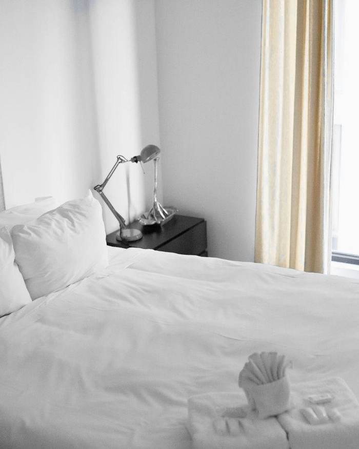 Cama blanca junto a una ventana, con lámpara de lectura y cortina clara