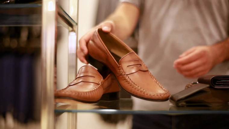Unas manos sostienen un mocasín de cuero junto a otro, sobre una repisa de vidrio
