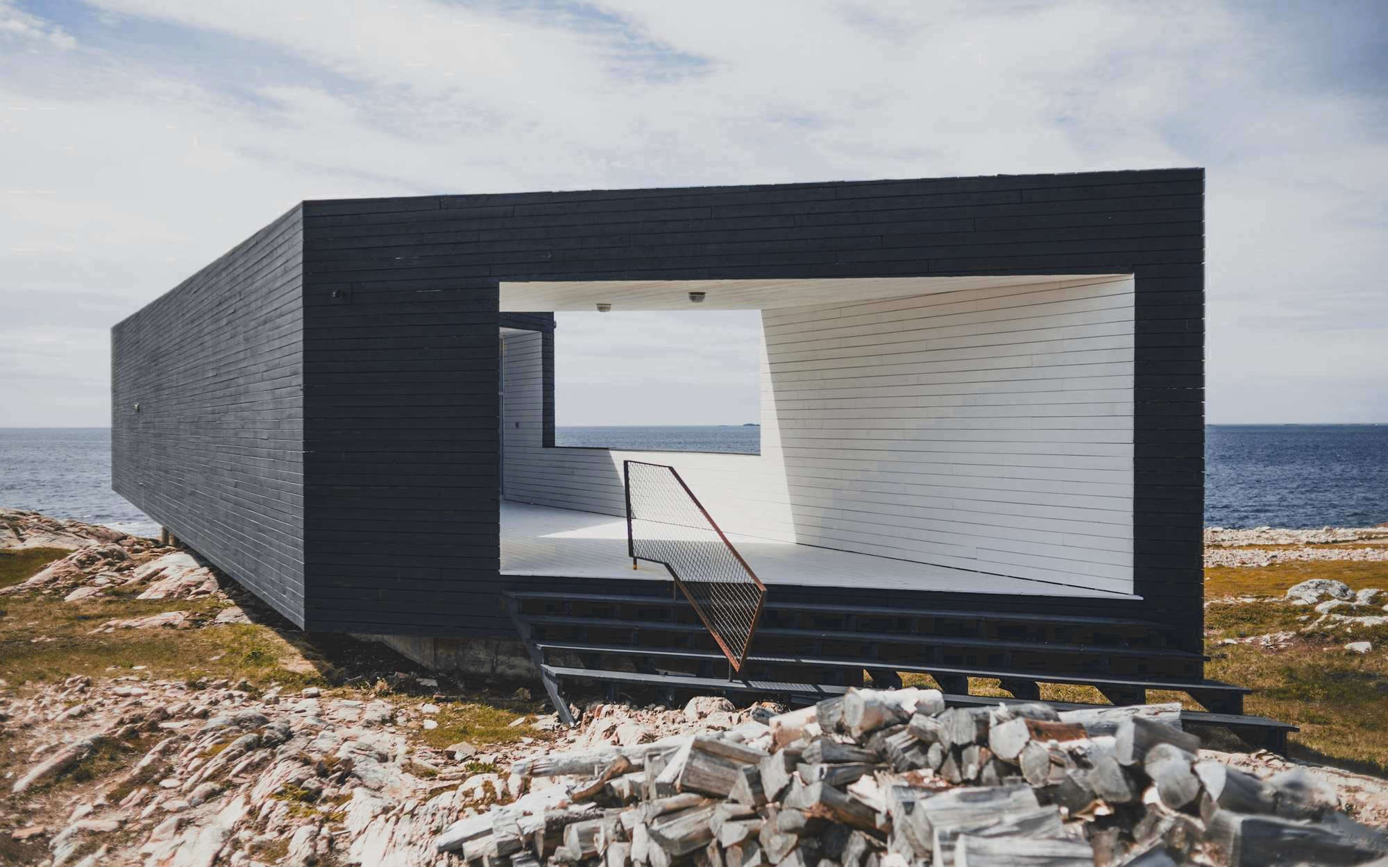 Casa negra en forma de caja abierta sobre una costa de rocas, con el mar al fondo