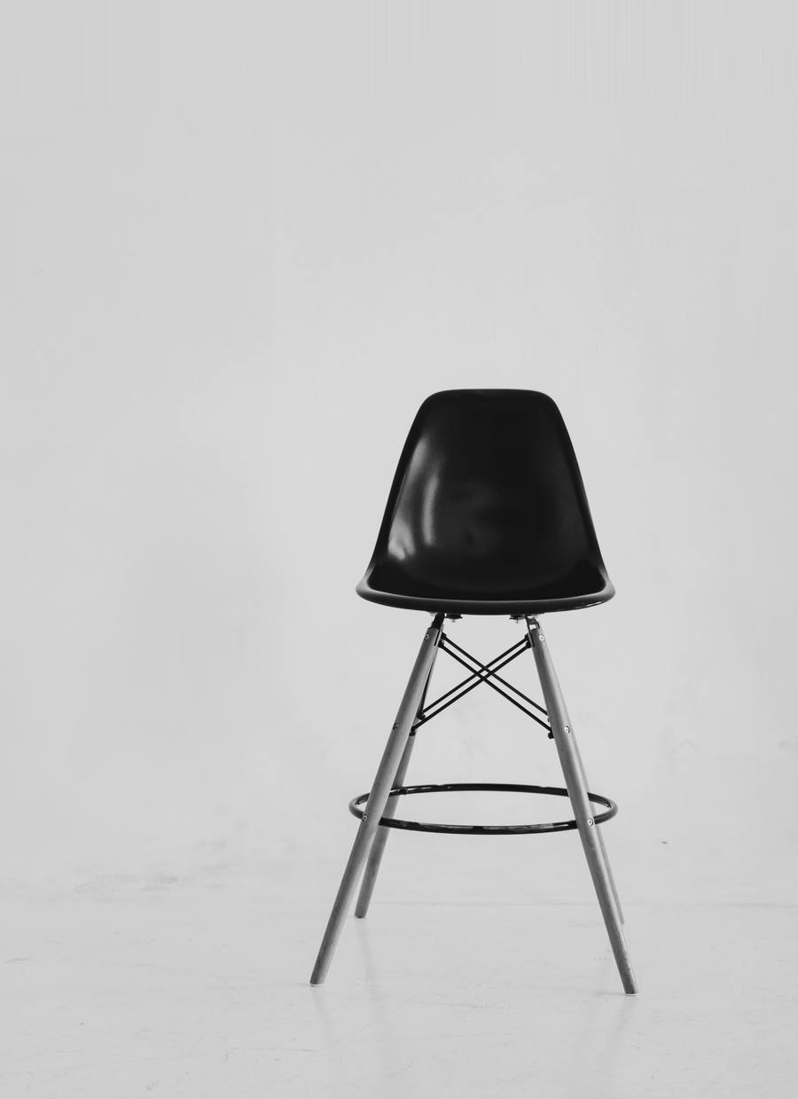 Silla de carcasa negra sobre patas de madera frente a un muro gris claro, en blanco y negro