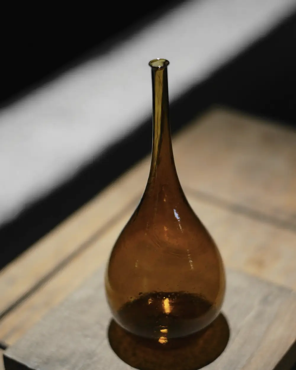 Botella de vidrio ámbar de cuello largo sobre una tabla de madera, con un haz de luz en diagonal