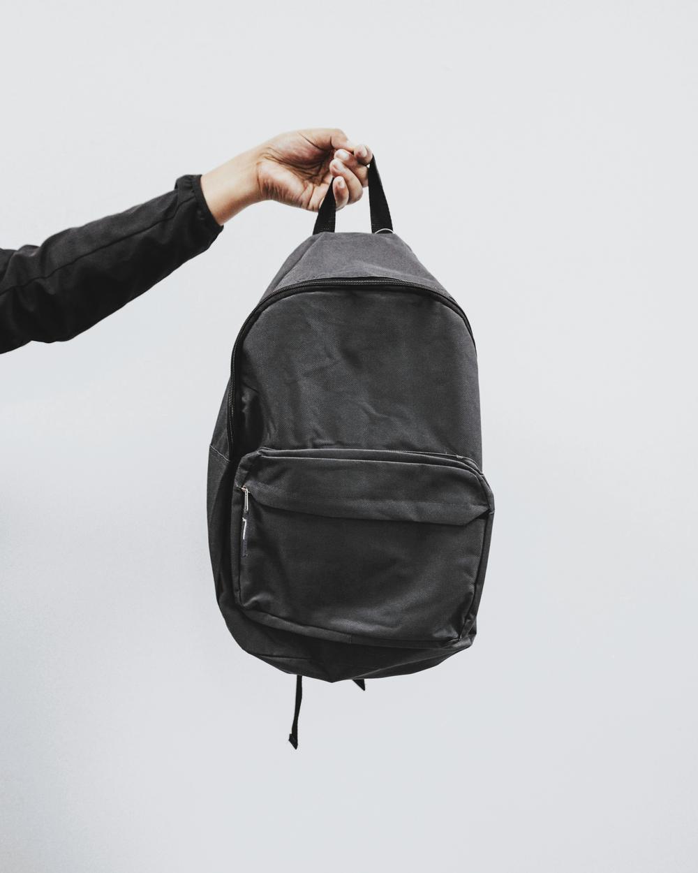 Una mano sostiene una mochila negra por su asa, contra un muro claro