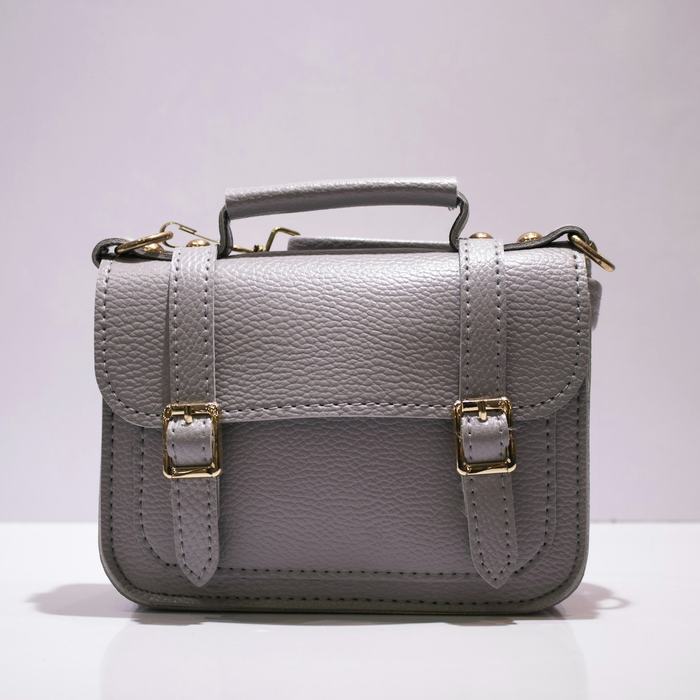 Bolso gris de solapa con dos hebillas, sobre un fondo claro