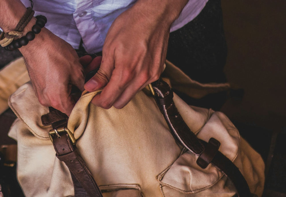 Dos manos abren un bolso de lona crema con hebillas de cuero