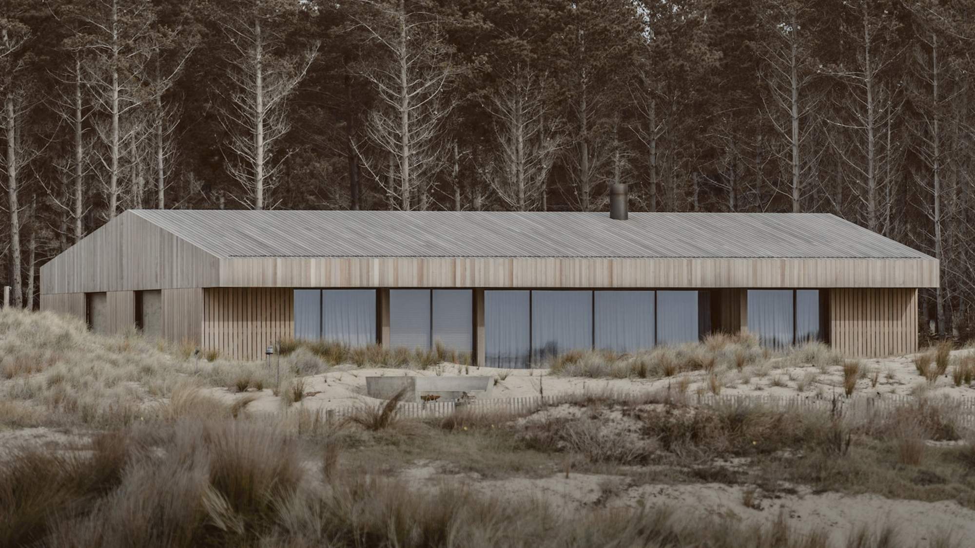 Una casa baja de madera clara y grandes ventanales, rodeada de dunas, con un pinar oscuro detrás
