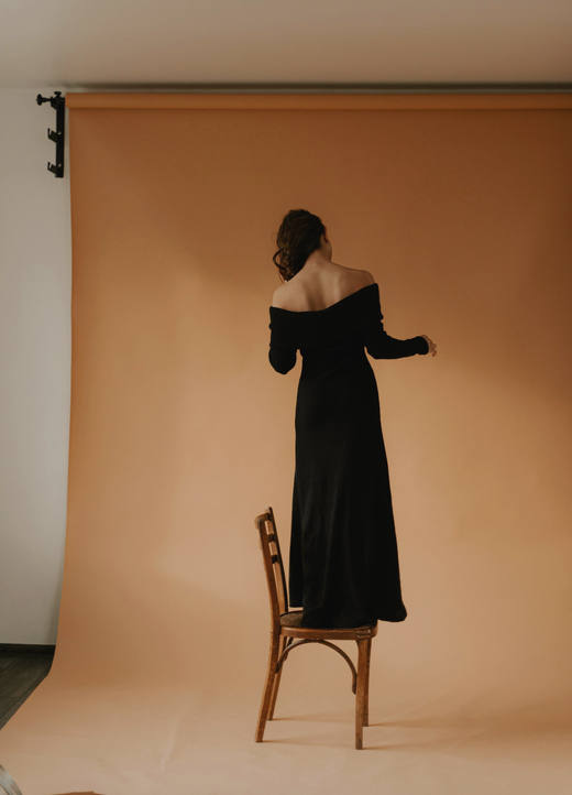 Figura de espaldas con un vestido negro de hombros caídos, subida a una silla frente a un fondo de papel ocre
