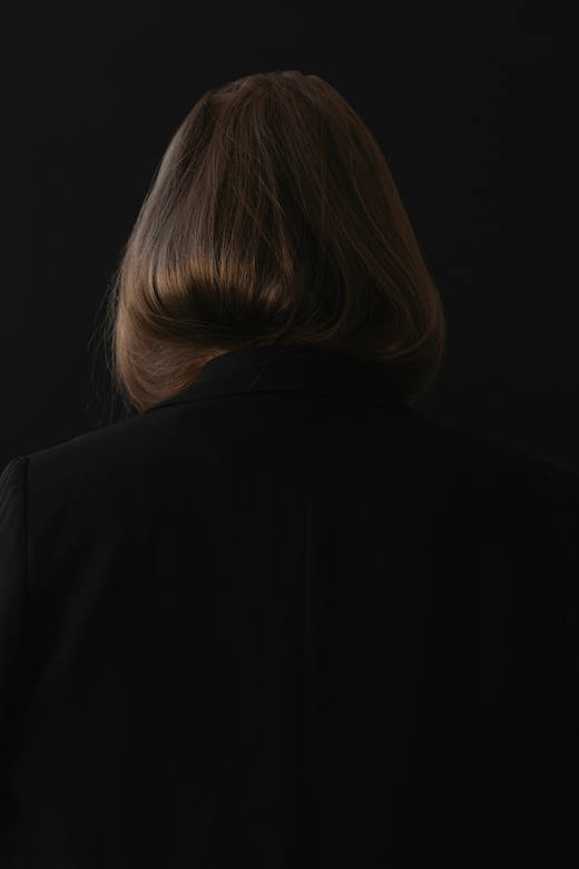Cabello castaño y abrigo negro, vistos de espaldas sobre un fondo oscuro