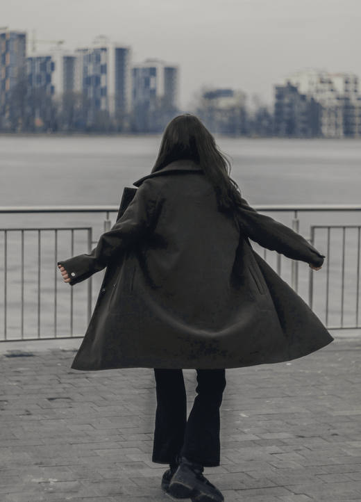 Persona de espaldas con un abrigo oscuro que vuela, frente a un paseo junto al agua