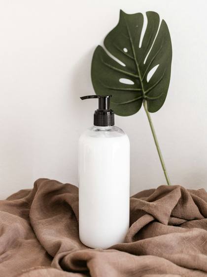 Botella blanca con dosificador junto a una hoja grande de monstera, sobre tela marrón