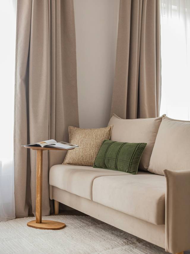 Sofá beige con cojines y una mesita auxiliar de madera junto a una cortina de lino