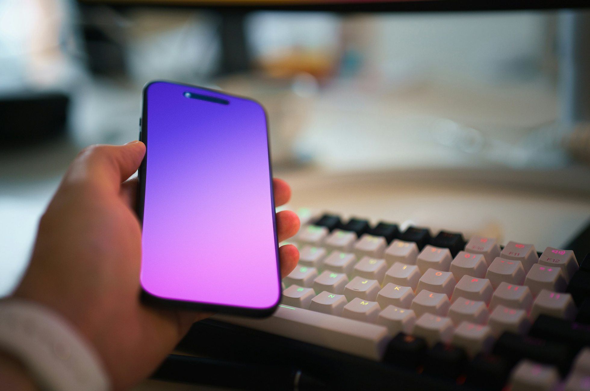 Una mano sostiene un teléfono con una pantalla de degradado violeta junto a un teclado mecánico