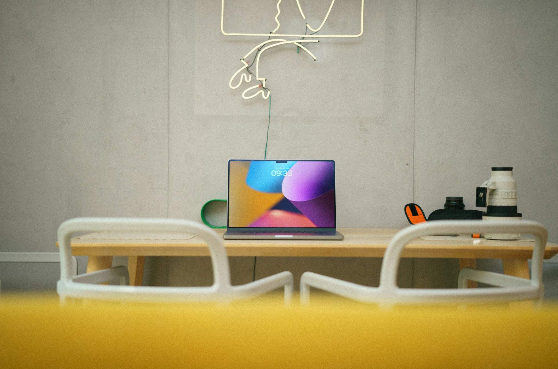 Un computador portátil sobre una mesa de madera frente a un muro de hormigón con un letrero de neón y sillas blancas