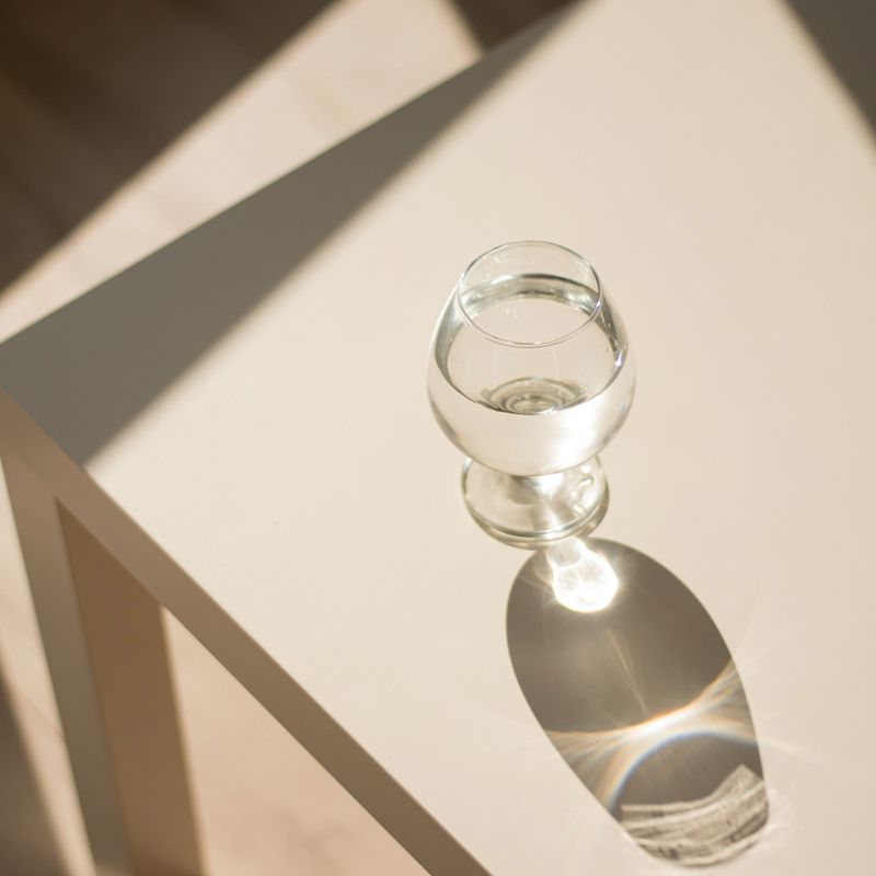 Copa de vidrio sobre una mesa blanca, con su sombra y un destello de luz