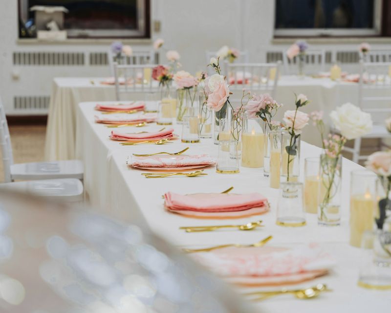 Mesa larga con mantel blanco, servilletas rosadas, rosas en vasos y velas de cera claras