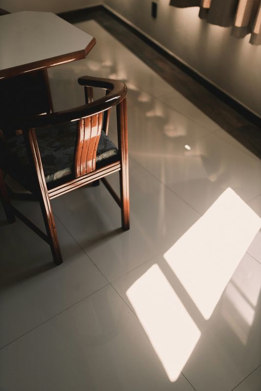 Silla de madera junto a una mesa, con rectángulos de sol sobre un piso brillante