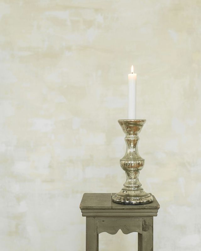 Vela blanca encendida en un candelero plateado, sobre un pedestal de madera y un muro de cal