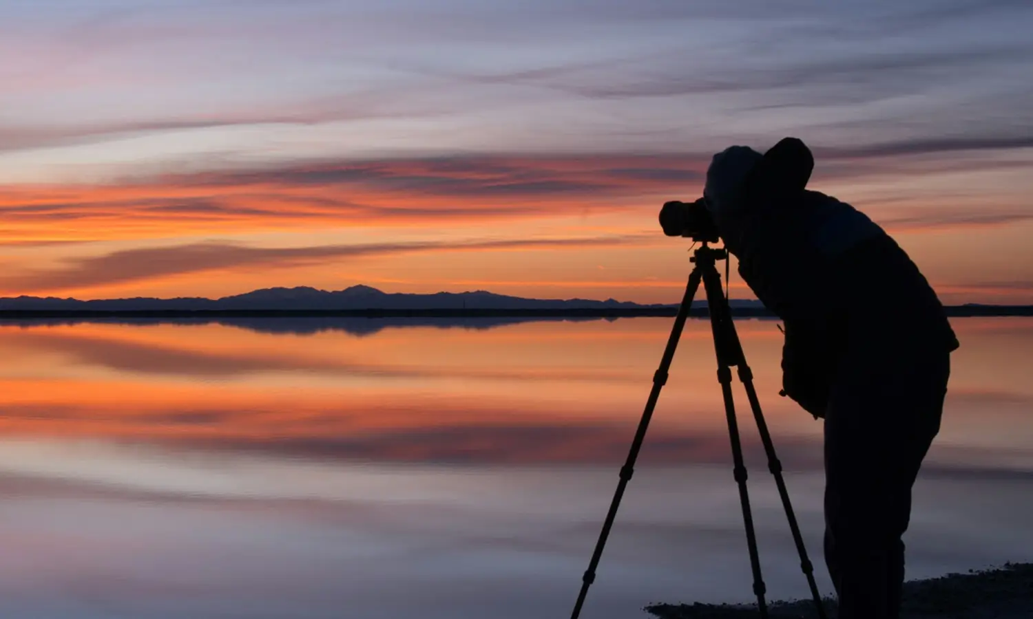 Silueta de un fotógrafo con trípode frente a un lago al atardecer