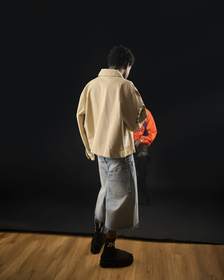 Figura de espaldas con chaqueta crema sobre un escenario oscuro
