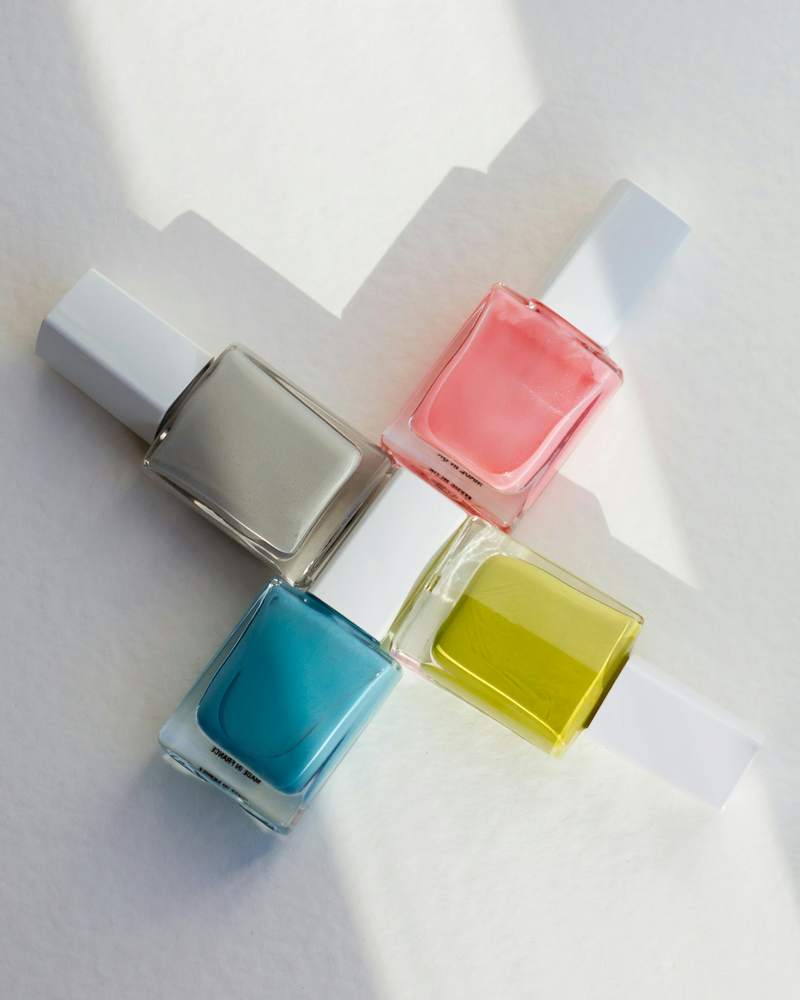 Cuatro frascos de esmalte en gris, rosa, turquesa y amarillo, cruzados sobre papel blanco