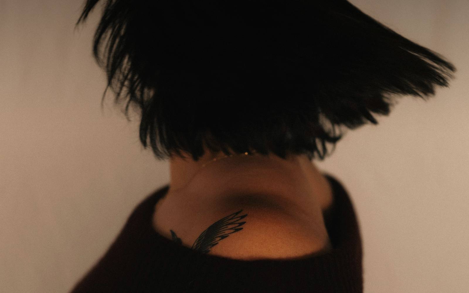 Una mujer de espaldas con el pelo corto en movimiento sobre un fondo color arena, vestida de negro