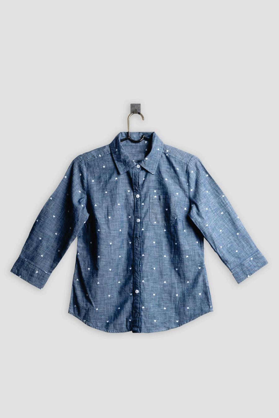 Una camisa de mezclilla azul con corazones blancos colgada de un gancho sobre un fondo gris