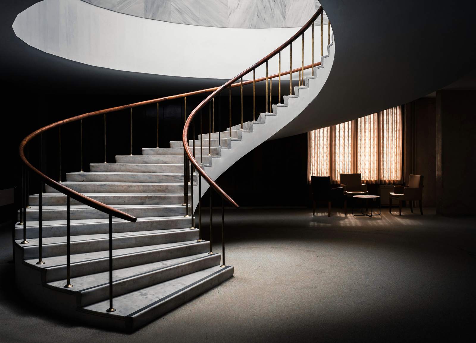 Escalera curva de peldaños claros con baranda de bronce y balaustres negros, en un vestíbulo oscuro