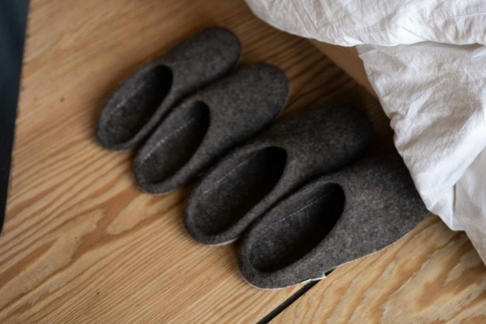 Pantuflas de fieltro gris oscuro junto a una cama con sábanas blancas