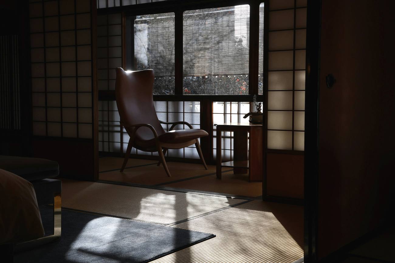 Sala en penumbra con una silla de cuero junto a una celosía de papel y una ventana por donde entra la luz