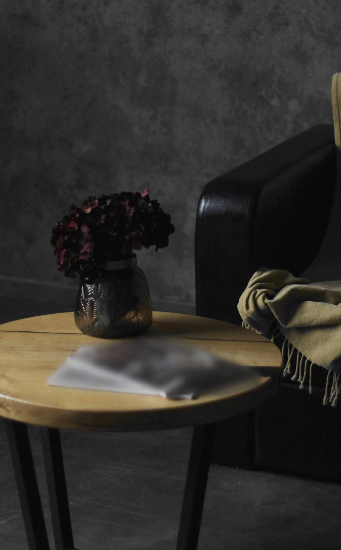 Rincón en penumbra: un sillón negro cubierto con una manta color arena y una mesa redonda de madera con flores secas en un florero
