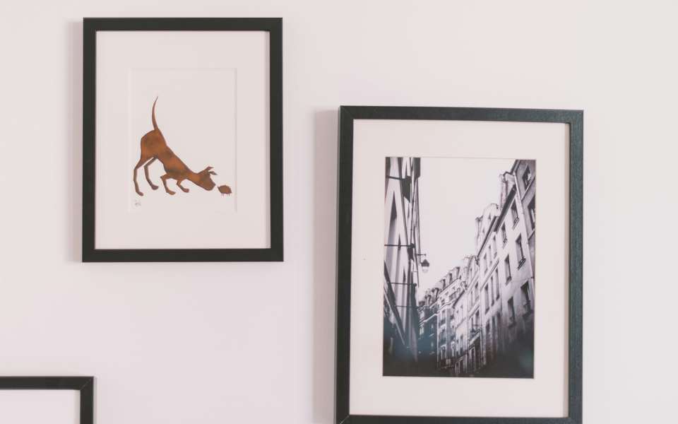 Dos láminas con marco negro sobre una pared rosada: un dibujo de un perro y una fotografía de edificios