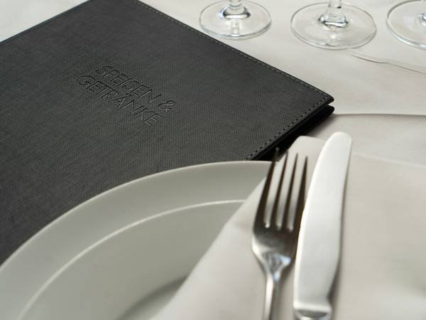 Carta de tapa negra y un plato blanco con cubiertos sobre un mantel claro